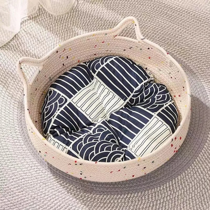 Panier chat rotin à deux poignées avec coussin gris à motifs blancs, posé sur un tapis gris